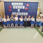 Grupa dzieci w białych koszulach i niebieskich spódniczkach stoi przed niebieską kurtyną. Napis Dzień Babci i Dziadka, czerwone serca. Dzieci są w sali z jasnymi ścianami i podłogą.