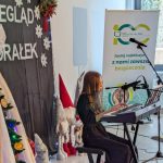 Na zdjęciu widać dziewczynkę grającą na keyboardzie, obok choinki z ozdobami na tle świątecznym. Tło ma dekoracje z gwiazdkami i wieńcem, a obok stoi baner z napisem. Pomieszczenie jest jasne z oknem po prawe