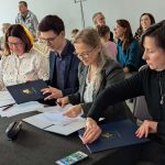 Na zdjęciu widać grupę dorosłych osób przy stole podpisujących dokumenty. Tło jest szare, a uczestnicy są ubrani w ciemne i jasne kolory. W tle siedzą inni ludzie, a na stole leży butelka wody i telefon.