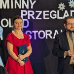 Na zdjęciu kobieta w czerwonej sukience i mężczyzna w garniturze stoją na scenie. Tło zdobią białe napisy i śnieżynki. Obok widoczna jest ozdobiona choinka.