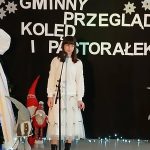 Dziewczyna w białej sukience śpiewa na tle czarnego plakatu z napisami. Obok przystrojona choinka i świąteczne krasnale. Tło ozdobione śnieżynkami, obok stojak z zielonym wieńcem.