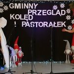 Scena ozdobiona świątecznie z choinką i figurkami gnomów. Kobieta w czerwonej sukience przemawia. Tło czarne z białym napisem.