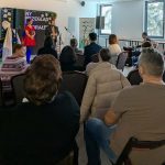 Na zdjęciu jest scena z występem w sali pełnej ludzi. Tło jest jasne z niebieskim światłem, choinką i krzyżem. Publiczność siedzi na ciemnych krzesłach.