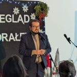 Na zdjęciu są dwie osoby na scenie, z mikrofonem i dekoracjami świątecznymi w tle. Kobieta w czerwonej sukience trzyma teczkę. Na czarnym tle są białe napisy i zawieszone śnieżynki.