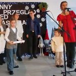 Grupa ludzi stoi na scenie z ozdobami świątecznymi na tle czarnego baneru z napisami. Kobieta w czerwonej sukience mówi do mikrofonu. Obok dzieci trzymające nagrody, na podłodze świąteczne dekoracje.