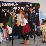 Grupa osób na scenie podczas przeglądu kolęd. Czarna dekoracja z napisem i śnieżynkami. Kobieta w czerwonej sukni czyta z kartki.