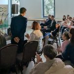 Na zdjęciu kobieta w czerwonej sukience przemawia do publiczności. Tło sceny jest ciemne z białymi napisami. Widzowie siedzą na krzesłach w jasnym pomieszczeniu.