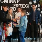 Na zdjęciu grupa osób na scenie, w tle czarne tło z dekoracjami świątecznymi. Dominuje kolor biały, czarny i czerwony. Chłopiec otrzymuje nagrodę.