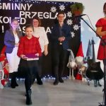 Na scenie stoi sześć osób, w tym dziecko w czerwonej koszuli, z mikrofonem w tle. Tło jest czarne z napisem i białymi dekoracjami. Kobieta po prawej trzyma teczkę.