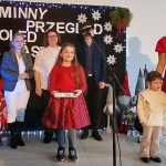 Grupa osób ubranych świątecznie stoi na scenie, tło czarne z napisem. Na scenie choinka z lampkami.