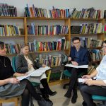 Cztery osoby siedzą w bibliotece, dyskutując. Tło to drewniane półki z kolorowymi książkami. Atmosfera jest skupiona i profesjonalna.