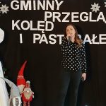 Scena z występem świątecznym, kobieta w czarnej bluzce w kropki śpiewa. Na tle napis Gminny Przegląd Kolęd i Pastorałek. Po lewej stronie dekoracje świąteczne i choinka.