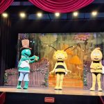 610695578_770323092756956_4064208452804774328_n Trzy postacie w strojach pszczół i świerszcza na scenie z kolorowym, kwiatowym tłem i czerwonymi kurtynami.