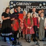 Grupa ludzi śpiewa na scenie z napisem Gminny Przegląd Kolęd. Dwie osoby grają na akordeonach. Tło jest jasne z niebieskimi i czerwonymi akcentami świetlnymi.