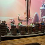 611016067_2062380997933867_8827817861985013037_n Grupa dzieci siedzi na atrakcjach przypominających statek w parku rozrywki. Tło to bajkowy krajobraz z różowym niebem i lodowymi górami. Na zdjęciu dominują ciepłe kolory i drewniane elementy.