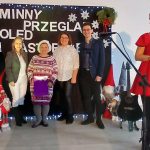 Grupa sześciu osób przed czarnym banerem z napisem GMINGNY PRZEGLĄD KOLED I PASTORAŁEK. Kobieta w czerwonej sukni z mikrofonem po prawej. Obok ozdobiona światełkami choinka.