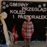 Na scenie świątecznej stoi osoba w zimowym stroju. Obok ozdobiona choinka z dużą kokardą. Tło z napisem i dekoracjami.