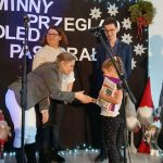 Grupa ludzi na scenie podczas wydarzenia bożonarodzeniowego. Przody, gitary, dekoracje w świątecznych kolorach. Dziecko odbiera nagrodę.