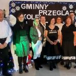 Grupa osób pozuje na tle ciemnego baneru z napisem Gminny Przegląd Kolęd. Ubrani w różne kolorowe stroje, stoją obok siebie. W tle ozdoby świąteczne i lampki.