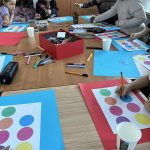 Grupa dzieci przy stole koloruje kółka na kartkach w kolorowe wzory. Na stole leżą flamastry, czerwone i niebieskie arkusze papieru. W tle znajdują się szafki i rośliny.