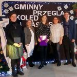 Zdjęcie przedstawia pięć osób na tle czarnego banera z białymi napisami i śnieżynkami. Po lewej stronie widoczne jest świąteczne drzewko z ozdobami. Tło ozdobiono kolorowymi światełkami i świątecznymi dekoracj