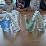 Dwa szklanki z niebieską i zieloną wodą połączone są ręcznikiem papierowym. W tle dzieci przy stole. Drewniane tło.