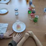 Dzieci przy stole z eksperymentem, kolorowe słoiki na brązowym tle, dwa talerze, z których jeden z odwróconą szklanką.