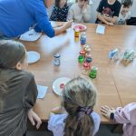 Dzieci siedzą przy drewnianym stole z kolorowymi słoikami i płynami. Osoba w niebieskiej bluzie wykonuje eksperyment. Tło jasne, okna w tle.