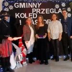 Grupa osób pozuje na tle ciemnej kurtyny z napisem GMINEY PRZEGLĄD KOLĘD I PASTORAŁEK. Dekoracje świąteczne, w tym śnieżynki i mikołaje. Jasne oświetlenie sceny, atmosfera uroczysto