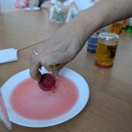 Ręka trzyma czerwoną świeczkę nad białym talerzem z różowym płynem. W tle stół z słoikami w różnych kolorach.