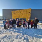 616491539_1928411838102027_102204658247170809_n Grupa dzieci w zimowych ubraniach stoi na śniegu przed szarym budynkiem z żółtym muralem. Po bokach znajdują się rzeźby na kolumnach. Czyste błękitne niebo w tle.