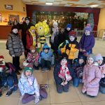 616927866_863123876338845_7522711784073896432_n Grupa dzieci pozuje z maskotką pszczółki w kolorowym wnętrzu. Wszystkie noszą zimowe ubrania. W tle widać kasy i mural z lasem.