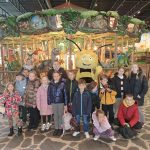 Grupa dzieci pozuje przed kolorową karuzelą z figurami dinozaurów. W tle dominuje bujna, zielona sceneria. Maskotka pszczoły towarzyszy dzieciom na pierwszym planie.