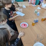 Grupa dzieci przy drewnianym stole, na którym stoją kolorowe szklanki i słoiki. Kilkoro dzieci eksperymentuje z płynami. Tło jest jasne, z widocznymi krzesłami i stołem.