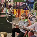 618580129_910903081464174_7187649311875408279_n Dziewczynka w szarej bluzie na różowym foteliku w wesołym miasteczku. Tło kolorowe z karuzelą. Jasne oświetlenie.