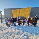 618808402_886149827628927_7794781116186076325_n Grupa dzieci stoi na śniegu przed szarym budynkiem z żółtym wrześniowym muralem. W tle bezchmurne, niebieskie niebo. Po lewej stronie widoczna rzeźba.