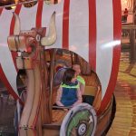 619063227_808580108895272_6793550282640988398_n Na zdjęciu są dwójka dzieci na atrapie statku Wikingów z białymi i czerwonymi żaglami. Statek jest na torze wodnym. Tło zbudowane w klimacie przygód, oświetlone ciepłymi kolorami.