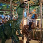619374301_1946820636209188_4243697739466850585_n Dziecko na karuzeli w kształcie dinozaurów, otoczone zielonymi liśćmi. W tle kolorowe malowidła z prehistorycznymi scenami. Jasne światła oświetlają atrakcję.