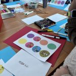 Na zdjęciu dzieci siedzą przy stole, rysują i kolorują. Na blacie znajdują się kolorowe kartki, kredki, pisaki. Tło jest jasne, widać pudełka i ścianę.