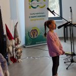 Dziewczynka w różowym swetrze śpiewa do mikrofonu. Obok stoją dekoracje świąteczne i roll-up z logo banku. Jasne wnętrze z oknem w tle.