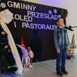 Chłopiec w dżinsowej kurtce stoi przed mikrofonem na tle sceny. Czarny baner z napisem Gminny Przegląd Kolęd i Pastorałek. Obok choinka i dekoracje świąteczne.