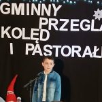Chłopiec w jasnej kurtce na scenie z czarnym tłem. Napis Gminny Przegląd Kolęd i Pastorałek. Dekoracje z śnieżynkami.