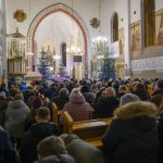Wnętrze kościoła z tłumem wiernych. Bogato zdobione ściany, choinki i żyrandole. Atmosfera świąteczna, kolory złote i białe.