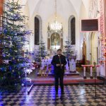 Wnętrze kościoła z choinkami ozdobionymi kolorowymi lampkami. Mężczyzna w garniturze stoi przy mikrofonie. W tle ołtarz i witraże.