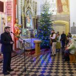 Mężczyzna przemawia do mikrofonu w kościele. Obok, przystrojona choinka i ludzie w tradycyjnych strojach. Tło zdobią dekoracje świąteczne i obrazy religijne.