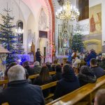 Wnętrze kościoła z ludźmi siedzącymi w ławkach. Bożonarodzeniowe dekoracje, kolorowe światła na choince. Na ścianach wiszą freski i obrazy religijne.