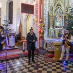 Kościół z dekoracjami i muzykami w ludowych strojach. Jasne ściany, kolorowe oświetlenie i choinka. Dwie osoby przy mikrofonach.