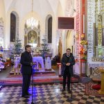 Wnętrze kościoła z dekoracjami świątecznymi, choinkami i ołtarzem. Na pierwszym planie stoją trzy osoby przy mikrofonach. Podłoga w czarno-białą szachownicę.