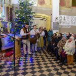 Wnętrze kościoła, mężczyzna przemawia przy mikrofonie, obok kolędujący muzykanci. W tle ozdobiona choinka i tłum słuchaczy. Kolory ciepłe i świąteczne.