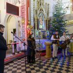 Wnętrze kościoła z chórem w strojach ludowych. Na tle choinki i ołtarza z kolorowymi dekoracjami. Posadzka w czarno-białą kratkę.